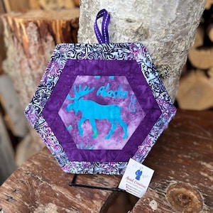 May include: Hexagon-shaped purple and blue quilted potholder with a teal moose silhouette and the word "Alaska". The potholder has a patterned border and a purple hanging loop. A small tag with text is attached.
