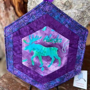 May include: Hexagonal purple and blue quilted potholder featuring a turquoise moose silhouette and the word "Alaska". It has a purple ribbon loop for hanging. The potholder is handmade.