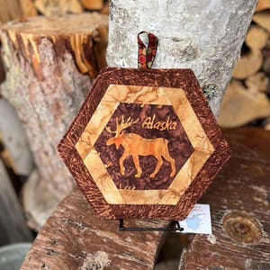 May include: Hexagon-shaped quilted potholder featuring a brown moose silhouette and the word "Alaska". The potholder has a brown and tan color scheme with a patterned border and a decorative hanging loop.