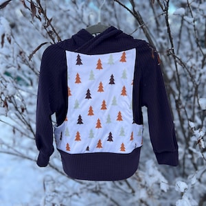 Puede incluir: Una sudadera con capucha de color púrpura oscuro con un panel blanco que presenta un patrón repetido de pinos naranjas, verdes y marrones. La sudadera está colgada de una percha sobre un fondo nevado borroso.