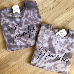 May include: Two grey and white tie-dye sweatshirts. One features the word "Thunder" in black script, the other "Monarchs". Each sweatshirt has a white tag with the letters "WH".