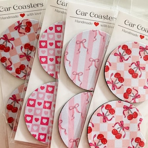 May include: Set of six pink and white car coasters with different patterns featuring cherries, bows, and hearts. The coasters are handmade and say "Car Coasters" and "Handmade with love".