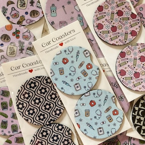 May include: A selection of car coasters with diverse patterns. Designs include floral motifs, medical icons, school items, and food illustrations. The packaging reads "Car Coasters Handmade with love".