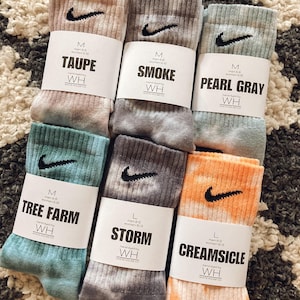 May include: Six pairs of tie-dye socks with different colour patterns and names. The socks are all white with different coloured dye patterns. The names of the socks are: Taupe, Smoke, Pearl Grey, Tree Farm, Storm, and Creamsicle. The socks are all size M or L.