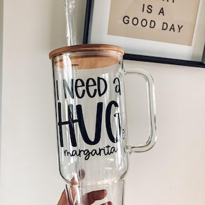 May include: A clear glass margarita mug with a wooden lid and a clear straw. The mug has a handle and the words "I need a HUG margarita" in black lettering. The background includes a framed print that says "Today is a good day".