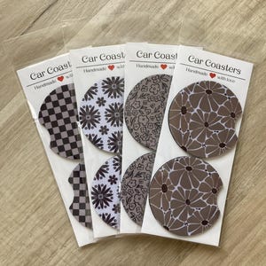 May include: Four sets of car coasters with different patterns. Each set includes two coasters. The patterns include black and white checkered, white with black floral, brown with black floral, and brown with white floral. The coasters are packaged in clear plastic bags with a label that says "Car Coasters" and "Handmade with love".