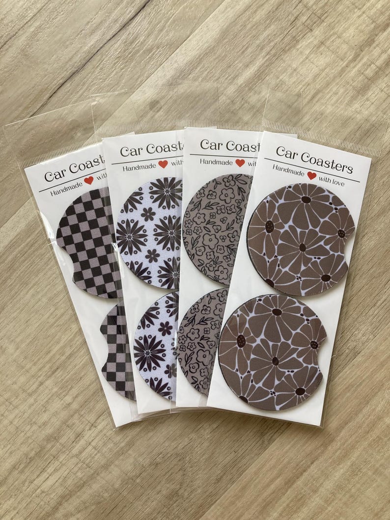 May include: Four sets of car coasters with different patterns. Each set includes two coasters. The patterns include black and white checkered, white with black floral, brown with black floral, and brown with white floral. The coasters are packaged in clear plastic bags with a label that says "Car Coasters" and "Handmade with love".