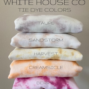 May include: A stack of five tie-dye sweatshirts in different colours, including taupe, sandstorm, harvest, creamsicle, and berry. The sweatshirts are folded and stacked on top of each other. The text "WHITE HOUSE CO TIE DYE COLORS" is at the top of the image.