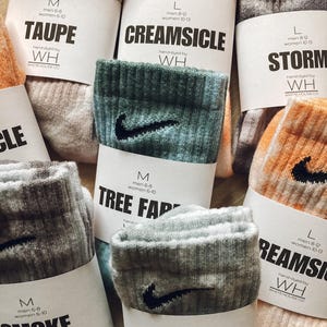 May include: Assortment of colourful socks with a Nike swoosh logo. Each sock has a label with a different colour name and size information. The colours include Taupe, Creamsicle, Storm, Tree Farm, Smoke, and Pearl Grey.