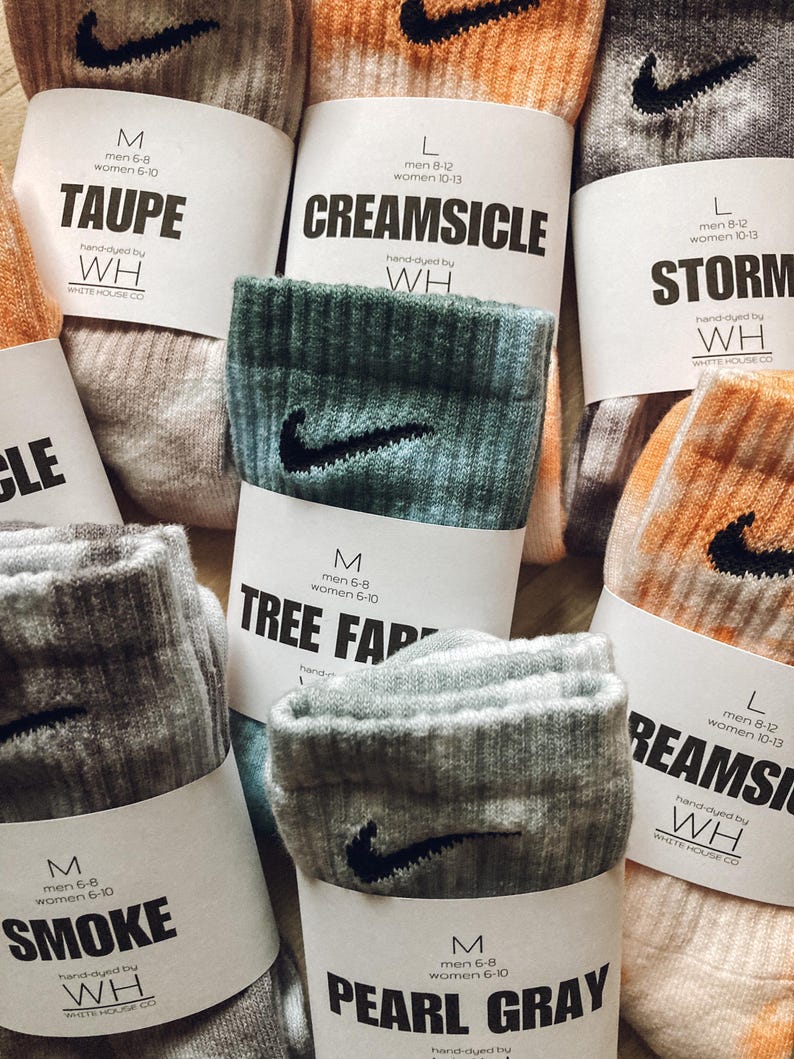 May include: Assortment of colourful socks with a Nike swoosh logo. Each sock has a label with a different colour name and size information. The colours include Taupe, Creamsicle, Storm, Tree Farm, Smoke, and Pearl Grey.