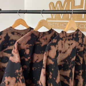 May include: Three brown and black tie-dye sweatshirts hang on wooden hangers from a black metal rod. The sweatshirts have a crew neck and a bleached pattern. A wooden sign with the text "WHITE HOUSE CO" is in the background.