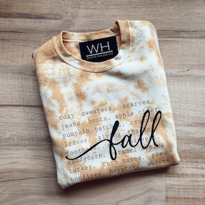 May include: A tan and white tie-dye crewneck sweatshirt featuring a black label with the text WH WHITE HOUSE CO at the neckline. The front displays a list of autumn-themed words in black typewriter font with the word fall in large script.