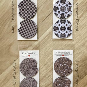 May include: Set of four car coasters featuring different floral and geometric designs. The coasters are packaged in clear plastic bags with labels that say "Car Coasters" and "Handmade with love". The designs include a mini checkered pattern, a mosaic floral pattern, a floral sketch pattern, and a large floral pattern.