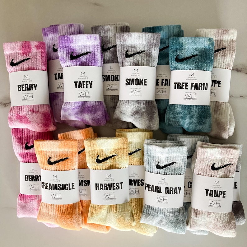 May include: A collection of 12 pairs of tie-dye socks featuring a Nike swoosh logo. The socks are in a variety of colours, including pink, purple, grey, blue, orange, and yellow. Each pair has a label with the size, colour, and name of the design.