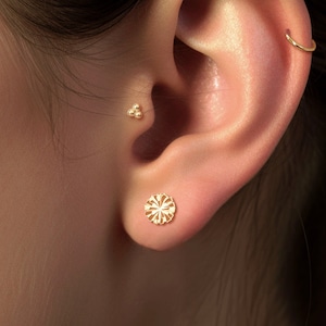 May include: Close-up of an ear adorned with gold jewelry. A small, delicate hoop earring is at the top, with a cluster stud and a floral stud earring on the lobe. The jewelry is set against a soft, neutral background.