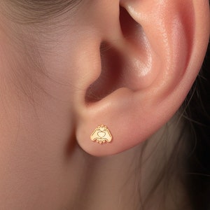 May include: Gold stud earrings featuring a small heart held between two hands.