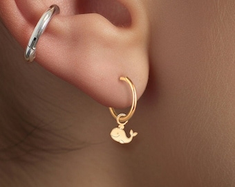 May include: A close-up of a person's ear with three gold earrings. The top earring is a small star with five points. The middle earring is a silver hoop. The bottom earring is a gold hoop with a small whale charm hanging from it.