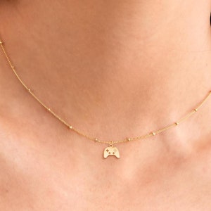 Puede incluir: Un collar de oro con un pequeño dije de oro en forma de mando de videojuego. El collar está hecho de pequeñas cuentas doradas.
