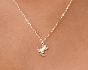 May include: A delicate gold necklace featuring a small frog pendant. The necklace is layered with a second, simpler chain. The frog pendant is detailed and adds a whimsical touch to the jewelry.