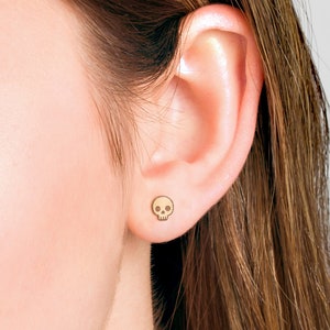Puede incluir: Primer plano de una oreja con un peque&ntilde;o pendiente de calavera de oro. El pendiente es un dise&ntilde;o sencillo con un contorno negro y ojos blancos.
