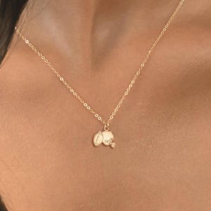 May include: A delicate gold necklace with two small charms. One charm is a small oval shape, and the other is a small flower shape. The necklace is worn around the neck.
