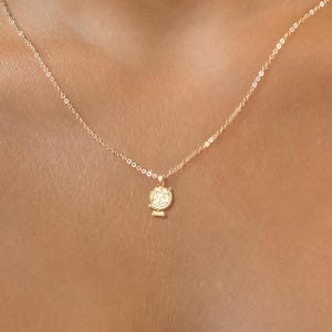 Puede incluir: Un delicado collar de oro con un pequeño y detallado colgante. El colgante parece ser un globo terráqueo o un diseño esférico similar. El collar se lleva alrededor del cuello, mostrando los intrincados detalles del colgante y el brillo sutil de la cadena.
