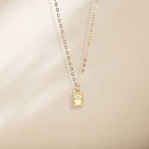 May include: A delicate gold necklace featuring a small, bear-shaped pendant. The pendant has a cream-colored center and a gold outline. The chain is also gold and has a subtle, linked design. The necklace is displayed against a neutral background.