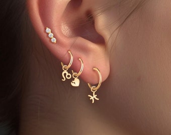 May include: A close-up of a person's ear with multiple gold hoop earrings. The earrings feature a heart charm, a star charm, and a zodiac sign charm. The person also has a small row of three diamond studs in their upper ear.