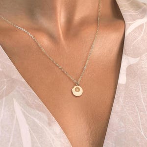 May include: A fine gold necklace featuring a small, circular pendant. The pendant has a minimalist design. The necklace is set against a patterned, light-coloured top, creating a simple and elegant aesthetic. The necklace is a delicate piece of jewellery.