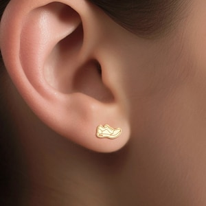 Puede incluir: Pendientes de botón de oro en forma de zapato. Los pendientes son pequeños y delicados, y presentan un diseño sencillo.