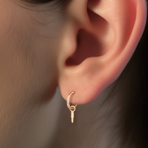 May include: A close-up of a person's ear with two gold earrings. The lower earring is a hoop with a small gold charm hanging from it. The upper earring is a small gold ball.