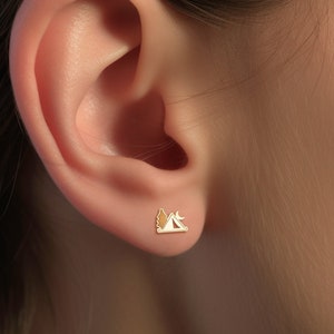 Puede incluir: Pendiente de oro con un pequeño diseño de escena de camping. El diseño presenta una tienda de campaña, un árbol y una luna creciente.