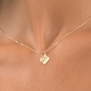 May include: A fine gold necklace with a small pendant. The pendant has a stylized design, possibly a crown or similar emblem. The necklace is displayed against a skin tone background.