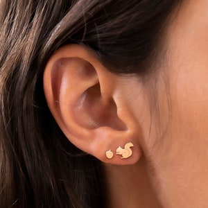May include: A pair of gold earrings featuring a small acorn and a larger squirrel design. The earrings are shown in a close-up view of a person's ear.