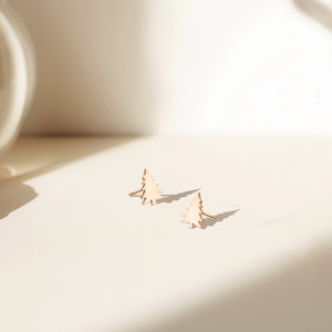 May include: A pair of small, light-colored wooden earrings in the shape of Christmas trees. The earrings are on a white surface, with shadows cast by sunlight. A vase and dried plant are in the background.