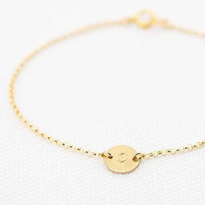 Könnte beinhalten: Ein zartes goldfarbenes Armband mit einem kleinen, runden Anhänger. Der Anhänger zeigt einen einzelnen Buchstaben, und das Armband besteht aus einer feinen Kette mit kleinen, runden Perlen. Der Verschluss ist ebenfalls goldfarben. Ein schlichtes und elegantes Schmuckstück.