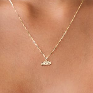 Può includere: Una collana dorata con due sottili catene. La catena inferiore presenta un piccolo ciondolo a forma di auto. La persona indossa un blazer bianco.