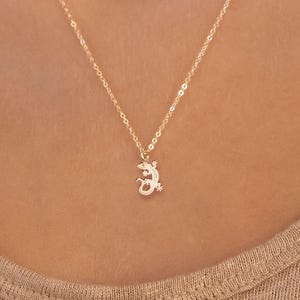 Op de afbeelding: Een delicate goudkleurige ketting met een kleine hagedis hanger. De hanger is ingewikkeld gedetailleerd en hangt aan een fijne ketting. De ketting wordt getoond tegen een neutrale achtergrond, wat het ontwerp van de sieraden benadrukt.