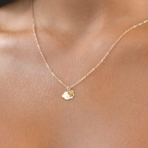 May include: Delicate gold-colored necklace with a small cloud pendant. The necklace features a thin chain and a charm shaped like a cloud with a small flower. The jewelry is worn around the neck.