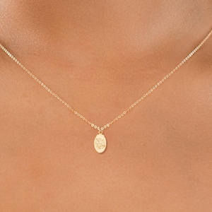 Könnte beinhalten: Eine zarte goldene Halskette mit einem kleinen, ovalen Anhänger. Der Anhänger hat ein dezentes eingraviertes Design und hängt an einer feinen Kette. Die Halskette wird über einem weißen Oberteil getragen.