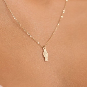 以下が含まれることがあります： 小さな、詳細なペンダントが付いた繊細なゴールドネックレス。ペンダントは、複雑なデザイン要素を持つ長方形の形をしています。ネックレスは人に見せられており、ジュエリーの配置とスタイルを強調しています。チェーンは細くてエレガントです。