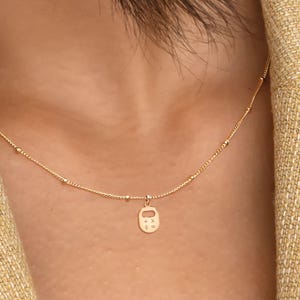 Puede incluir: Delicado collar dorado con un pequeño colgante con forma de casa. La fina cadena tiene pequeñas cuentas espaciadas uniformemente. El colgante está centrado y el collar se lleva sobre un fondo neutro.