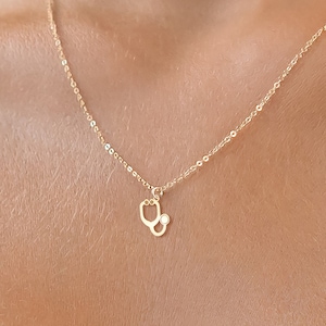 Op de afbeelding: Een gouden ketting met een klein stethoscoop-hanger met een enkele heldere steen.