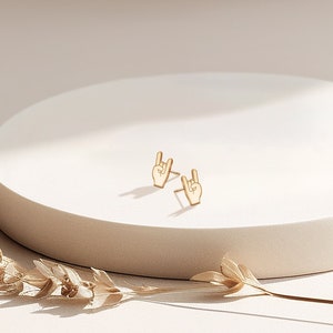 Puede incluir: Un par de pendientes de color dorado con un diseño de mano haciendo el gesto de rock and roll.