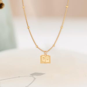 May include: A delicate gold necklace featuring a pendant shaped like an open book with a cross. The necklace has a fine chain with small, evenly spaced beads. The pendant and chain are gold-colored, creating a simple yet elegant design.