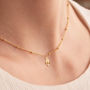 Könnte beinhalten: Eine goldene Kettenkette mit einem kleinen, zarten Anhänger in Form einer Hand. Die Kette besteht aus winzigen goldenen Perlen und wird von einer Person mit einem weißen Hemd getragen.