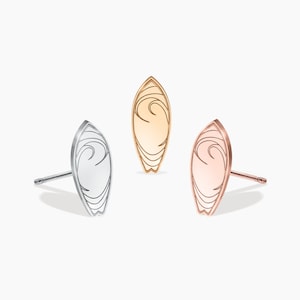 Puede incluir: Tres pendientes en forma de tabla de surf en plata, oro rosa y oro. Cada pendiente presenta un diseño de ola estilizado.