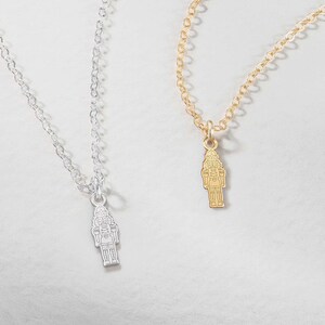 Puede incluir: Dos delicados collares, uno plateado y otro dorado, cada uno con un pequeño colgante en forma de cascanueces. Las cadenas son finas y los colgantes están detallados, sobre un fondo suave y neutro.