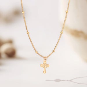 Op de afbeelding: Een delicate goudkleurige ketting met een kleine kruisvormige hanger. De ketting heeft een fijne ketting met gelijkmatig verdeelde kleine kralen. De kruishanger heeft een sierlijk ontwerp. De sieraden worden gepresenteerd tegen een zachte, vage achtergrond.