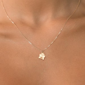 Puede incluir: Un collar de cadena de oro con un pequeño colgante de oro que representa una cara con las manos sobre los ojos. El collar lo lleva una persona con una blusa rosa claro.